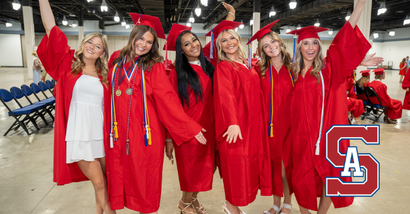Six high school graduates wearing red robes, posing side by side with their arms raised.  Behind them are more seated graduates . The setting is a large event center area.