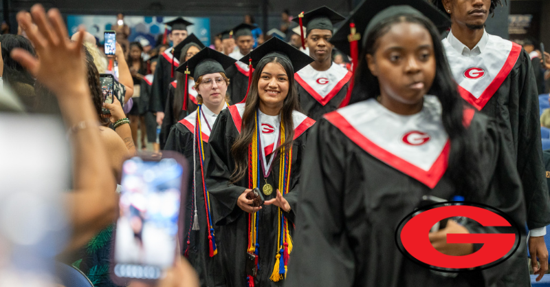 High school graduates wearing black robes with red accents walking down the middle aisle of a ceremony with families cheering them on