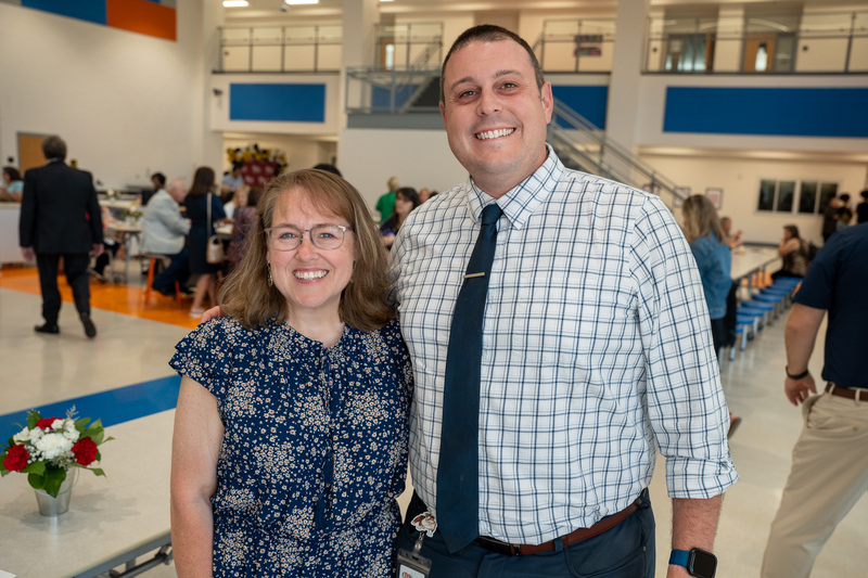 A principal wearing a plaid shirt and tie stands next to a retiree wearing a blue floral dress