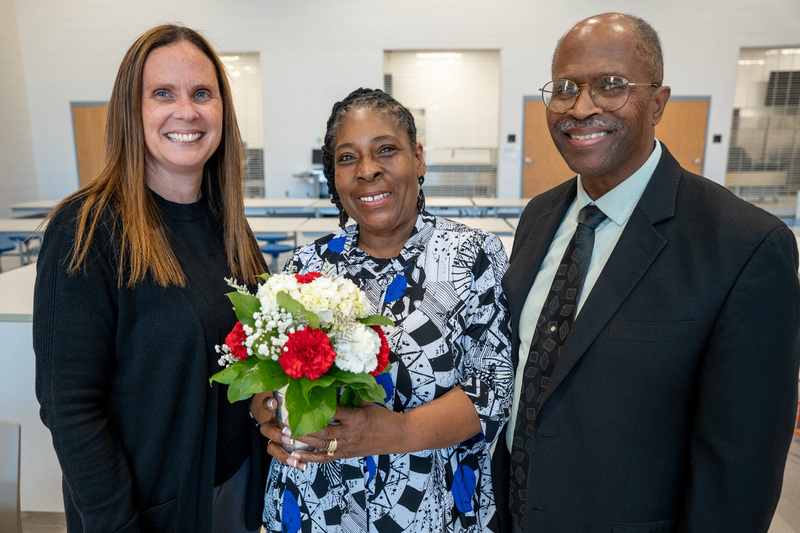 A retiree in a blue floral dress stands next to her husband and school principal, both wearing black formal attire