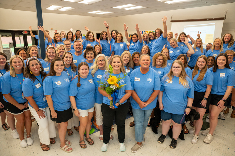 Large group of AO Elementary staff wearing blue shirts with Principal Erin Millspaugh in the center holding a flower bouquet