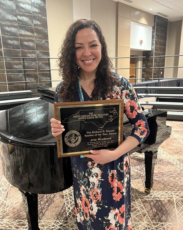 Aria Westbrook in a blue floral dress holding a recognition plaque at the NCMEA conference