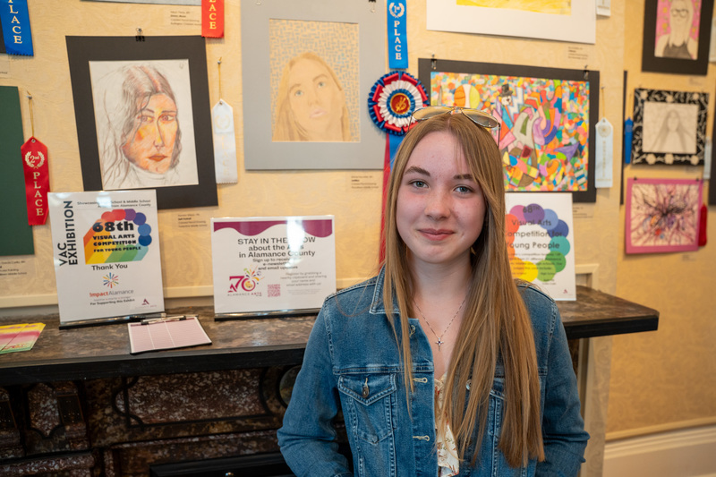 A teenage girl with long straight strawberry blonde hair and aviator glasses pushed up on her head smiles warmly at the camera in front of her First Place artwork.