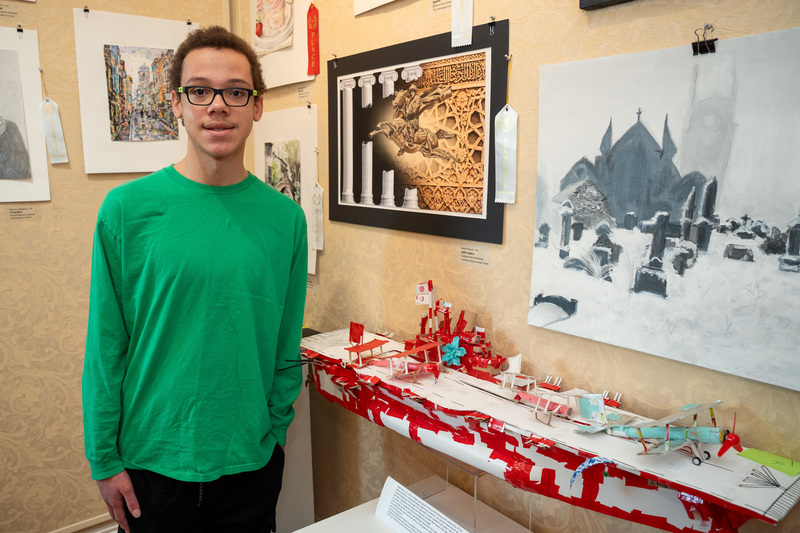 Western High student Dayne Wheeler standing in front of his large mixed-media ship art titled "Aircraft Carrier Ikigai" depicting the carrier in primarily red and white colors