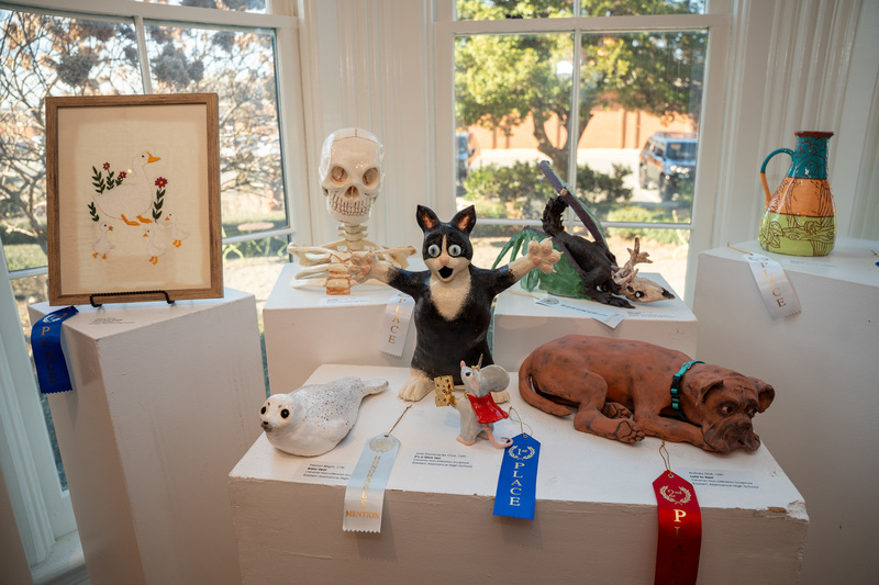 Several ceramic sculptures on display; a black cat surprised by a mouse king, a detailed skull and clavicular, a sleeping dog, a seal, and a pot
