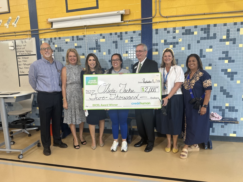 School and District leaders pose in gym with Coach Tacker holding oversized check for EXCEL Award