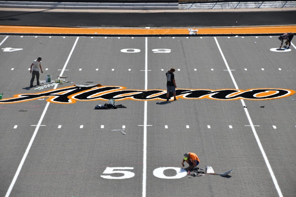 People on a gray turf football field with the words Alamo in black and gold in the center