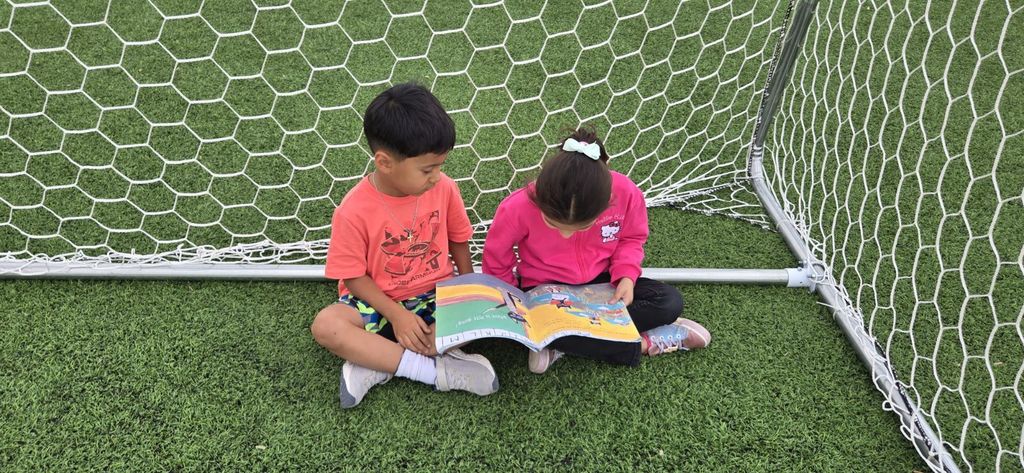 Two students sitting together on the turf in a soccer goal looking through a book
