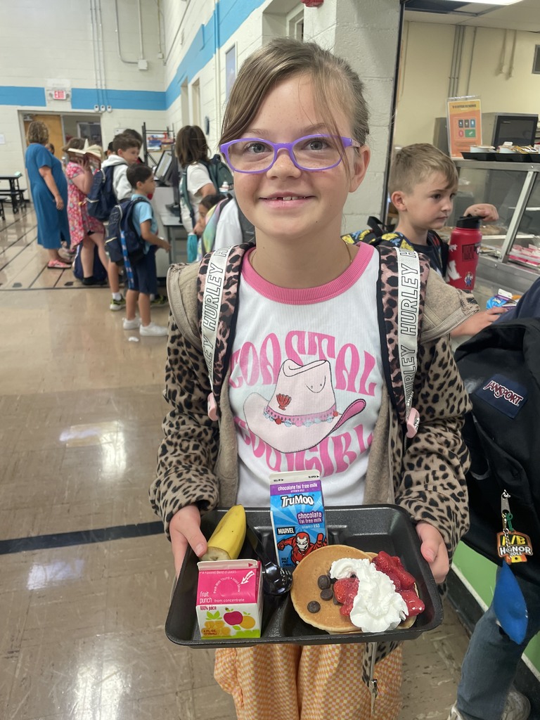 Student smiling holding breakfast tray with juice, milk, banana, and pancakes topped with strawberries and whipped cream