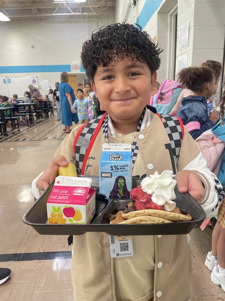 Student smiling holding breakfast tray with juice, milk, banana, and pancakes topped with strawberries and whipped cream