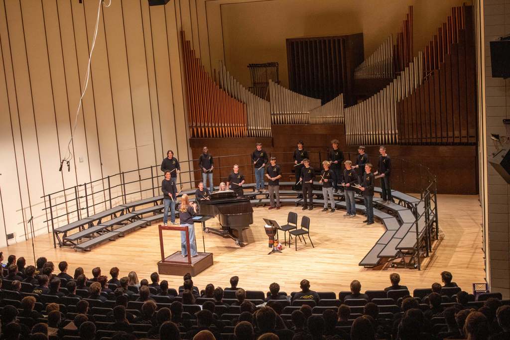 High school students standing on riser singing to audience while using drum sticks to play music