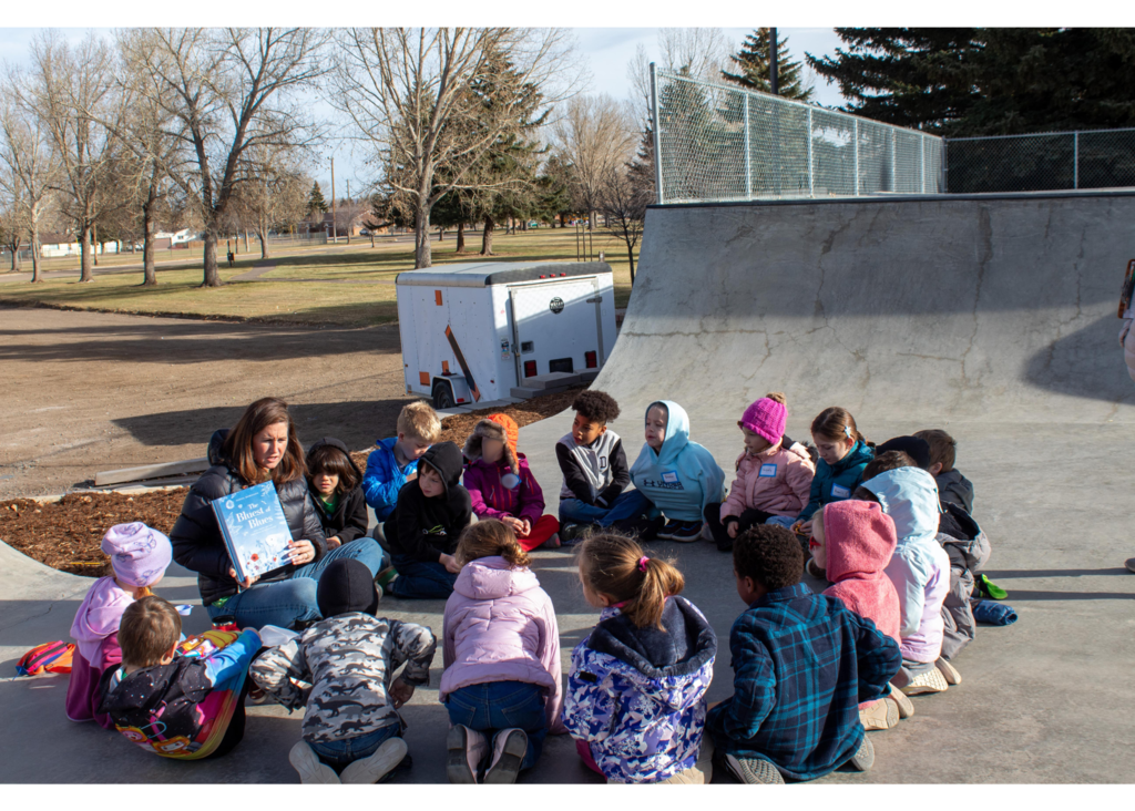 Students sitting in a circle outside being read a book by teacher