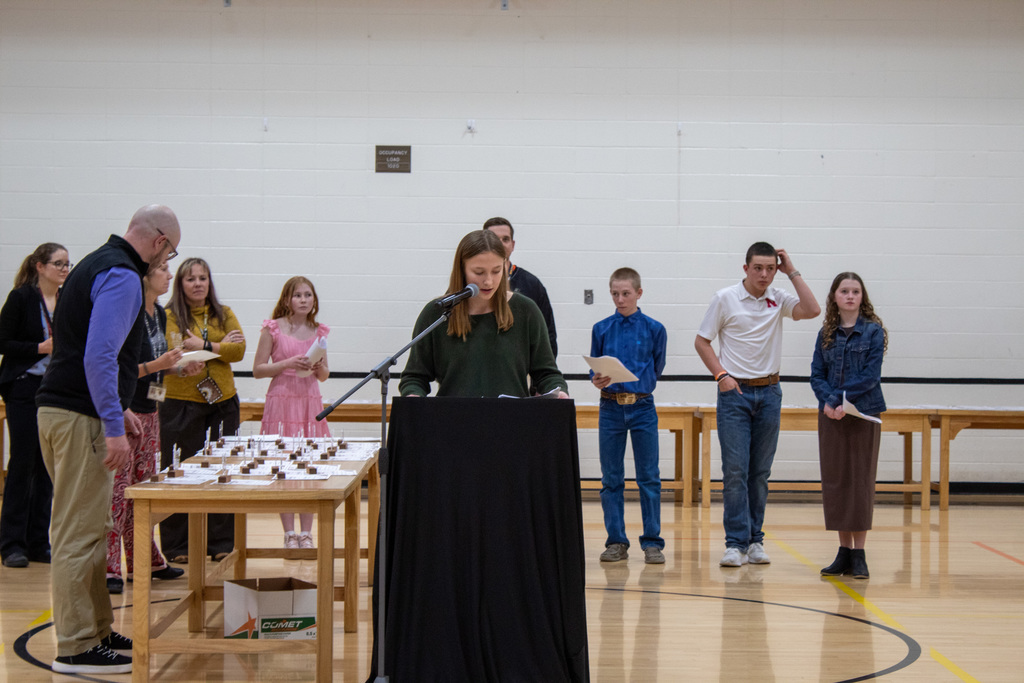 young female student with brown hair in standing at a podium surrounded by friends and teachers