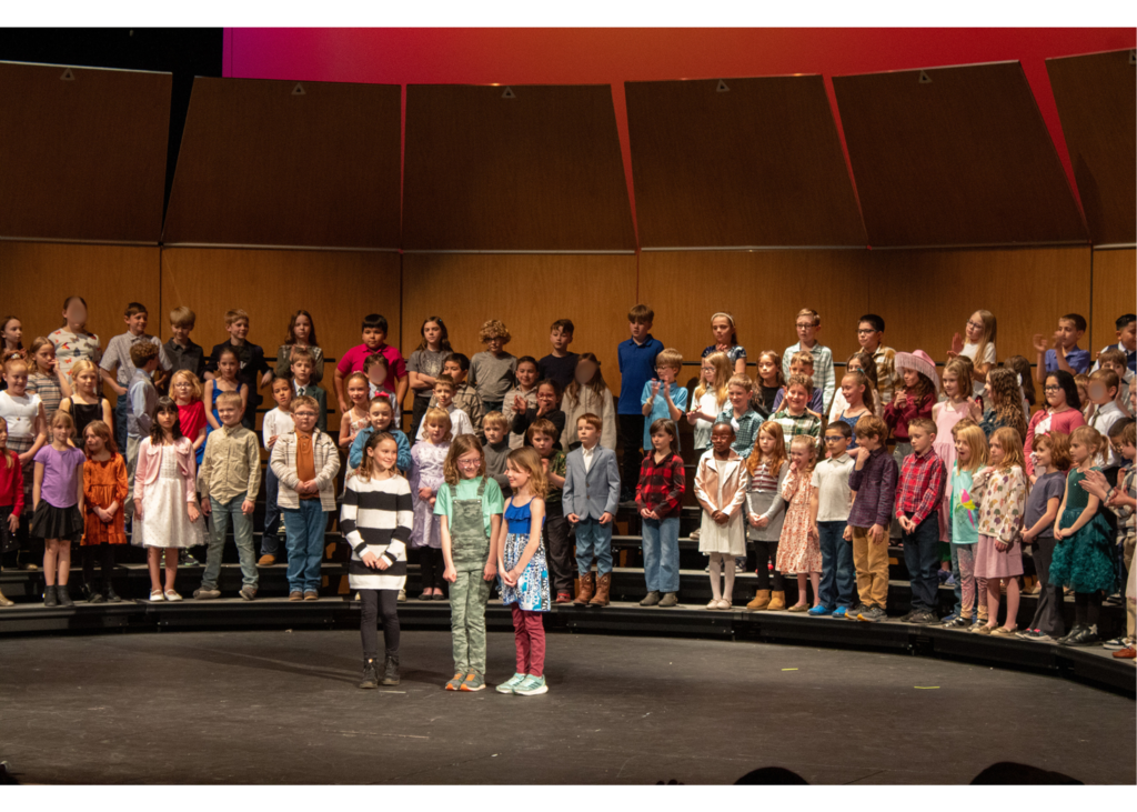 Three young girls standing in front of risers singing solo as the others watch