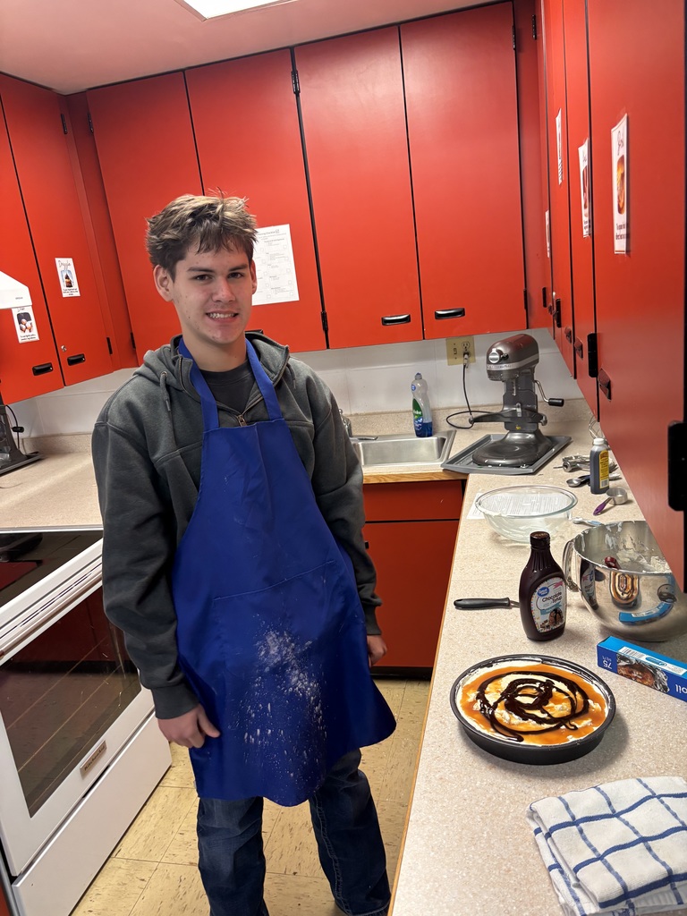 Male student in a blue apron standing next to a pie