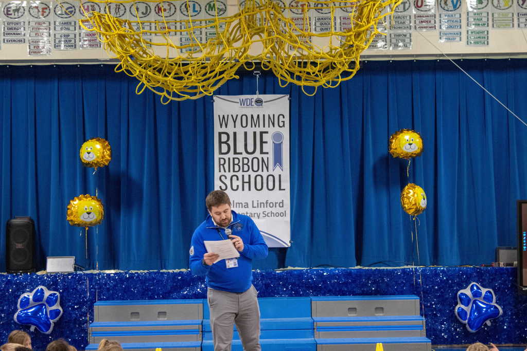 Man in blue shirt with dark short hair reading from a paper into a microphone