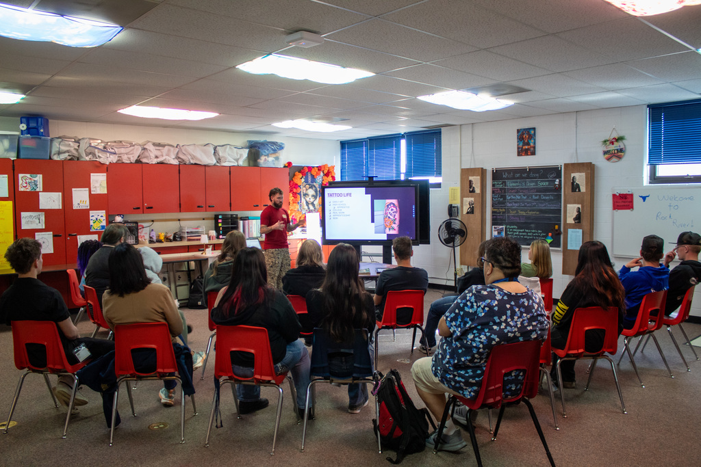 Classroom full of students sitting listening to a man talk at the front 