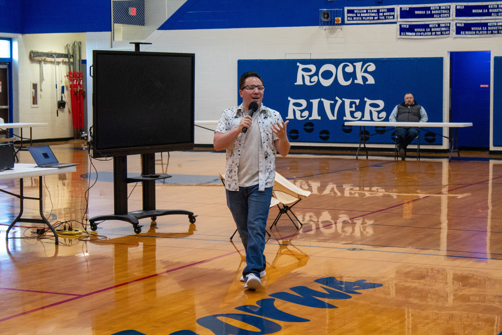 Man dressed in blue jeans and whit button down walking across a gym holding a microphone