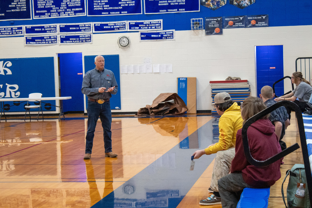 Man in jeans a blue shirt talking to students sitting in bleachers