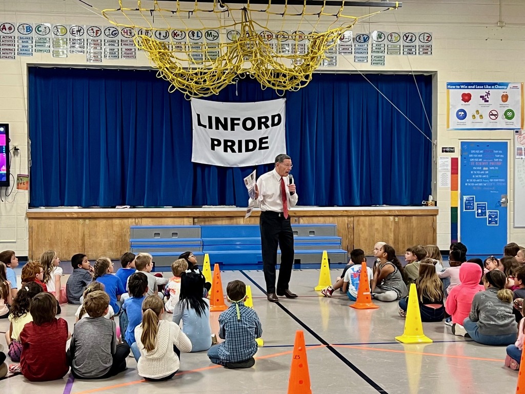 Man in white shirt and slacks talking to gym full of students 