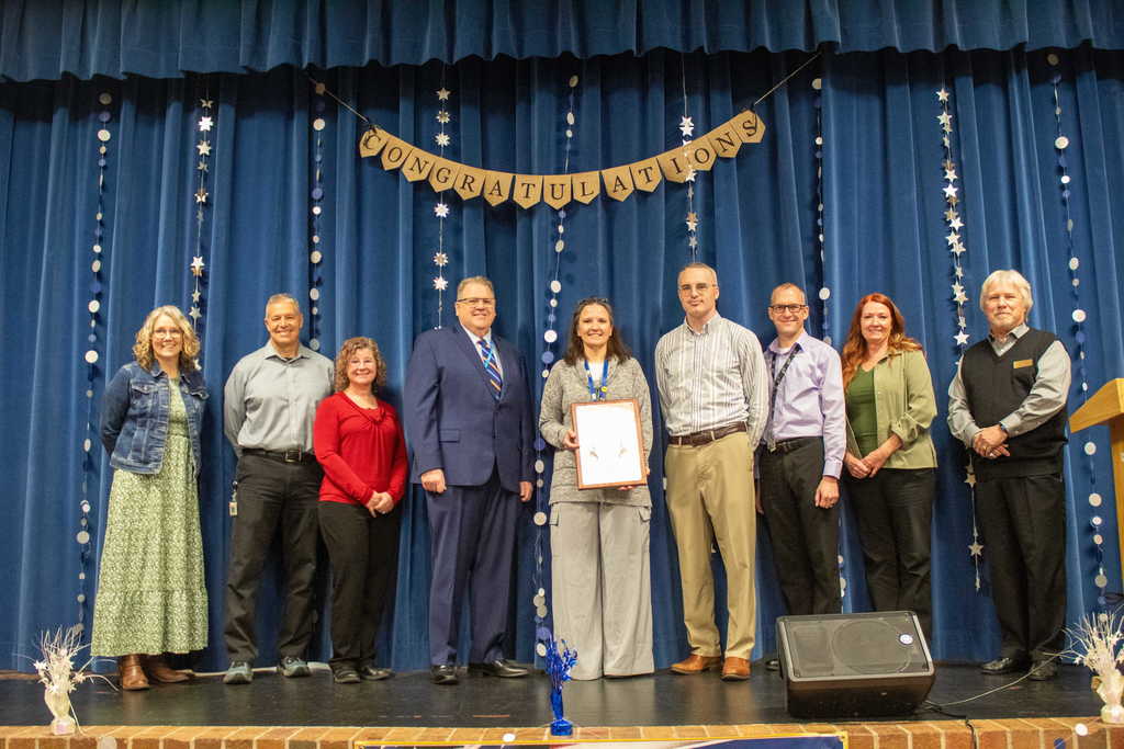 Group of adult standing on stage surrounding a women holding a plaque