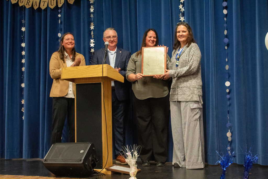 three adults standing on stage surrounding a women holding a plaque