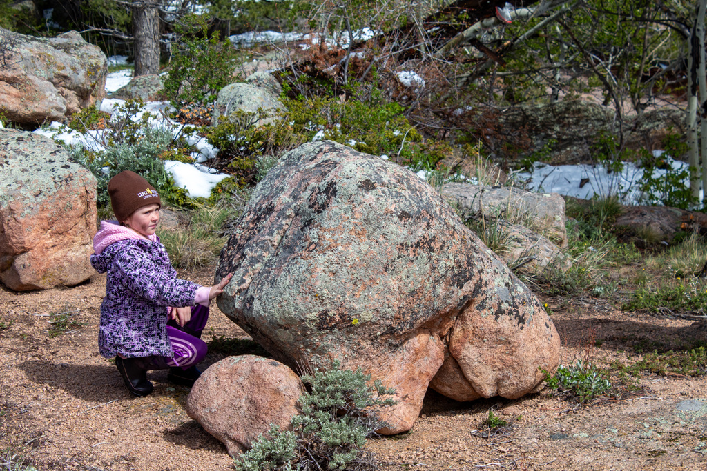 Young girl in purple coat and brown beaning leaning down by a rock