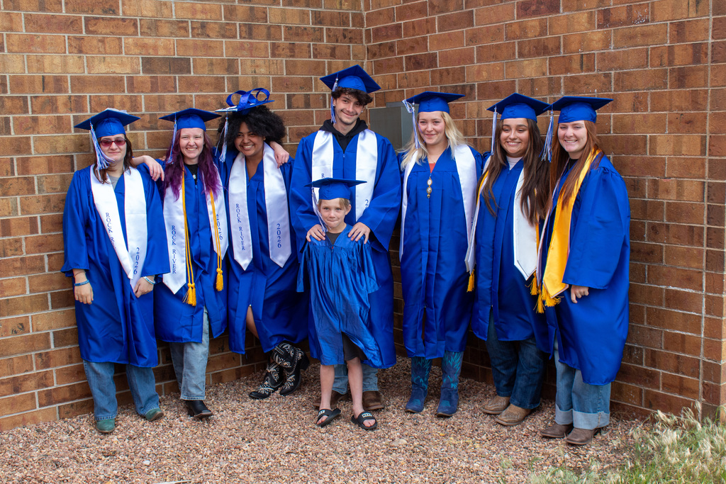 Group of seven students with blue graduation cap and gowns on standing with one younger boy dressed in a matching blue gown