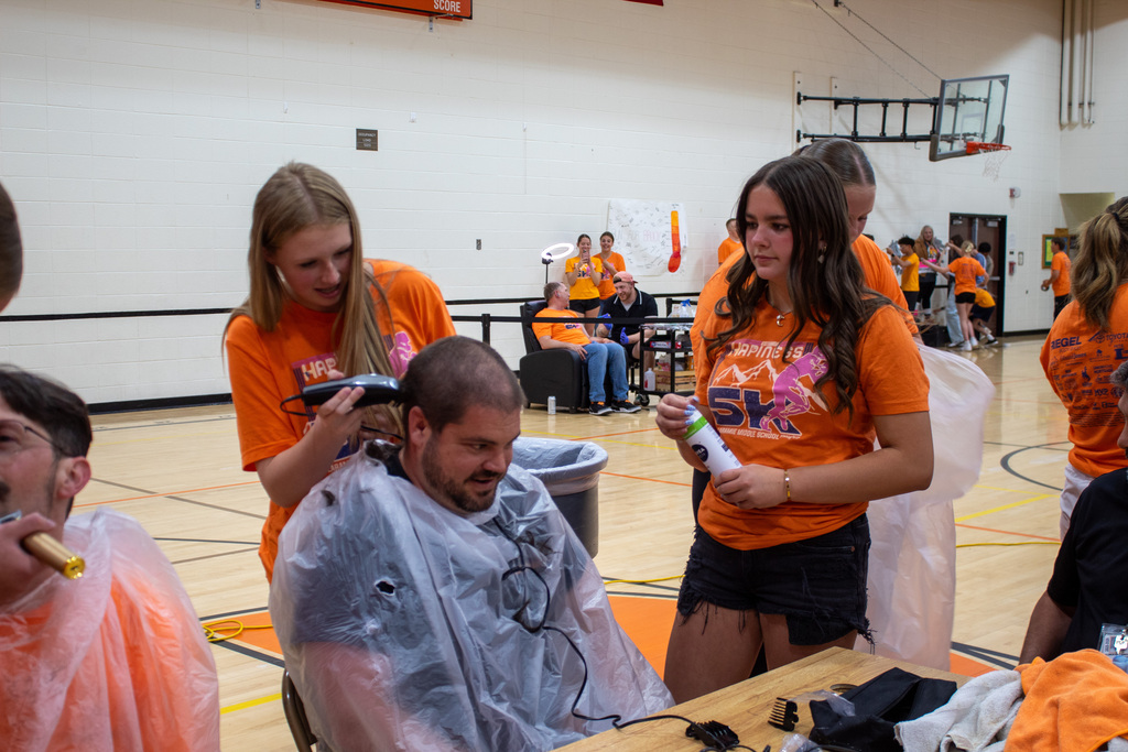 Two young girls in orange t-shirts shaving a mans head