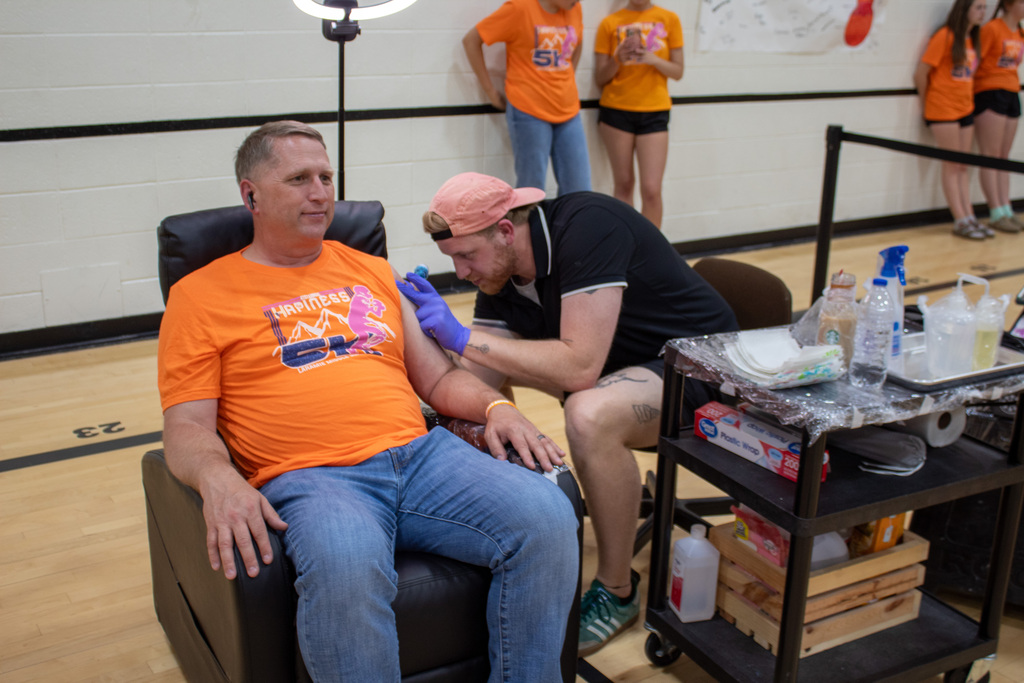 Man in orange shirt sitting on recliner getting a tattoo from a man dressed in black shorts and shirt