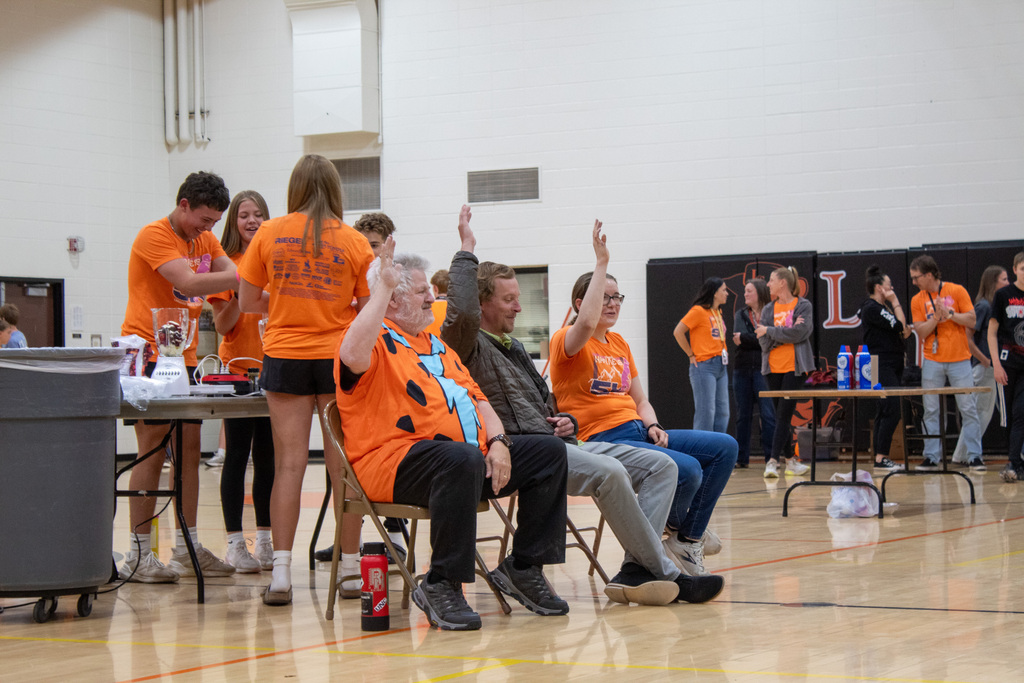 Three adults sitting in a row of chairs holding hands up while students stand behind them