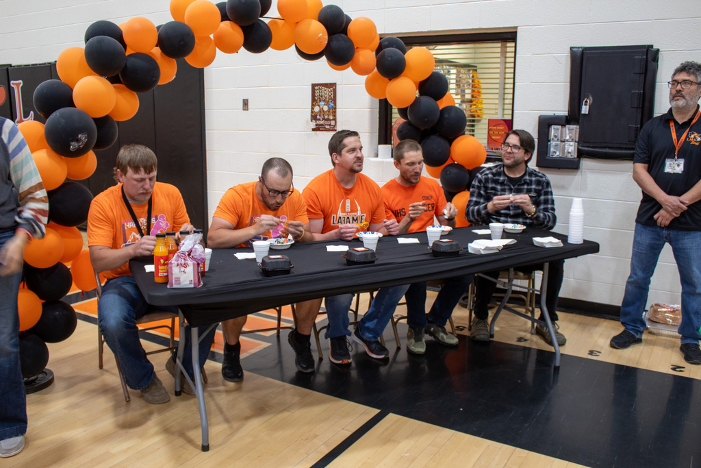 Four men in orange shirts sitting at a long table eating wings