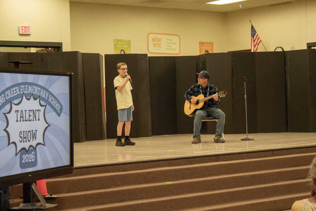 Young boy dressed in shorts and a shirt singing with teacher playing the guitar