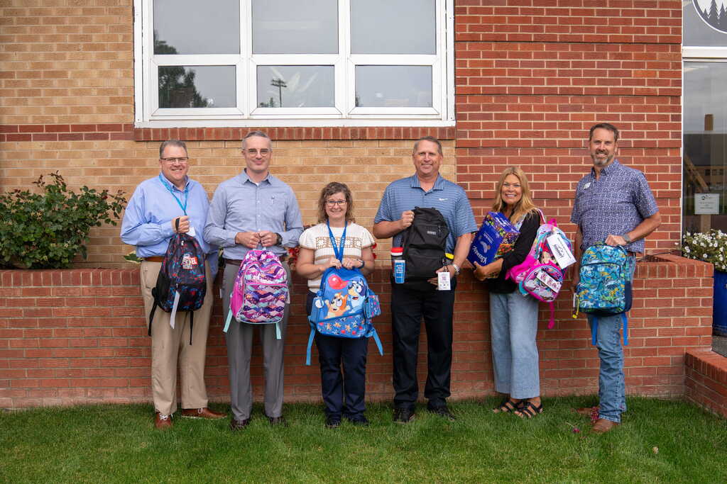 Group of men and women dressed in business clothes holding up childrens backpacks fulll of school supplies