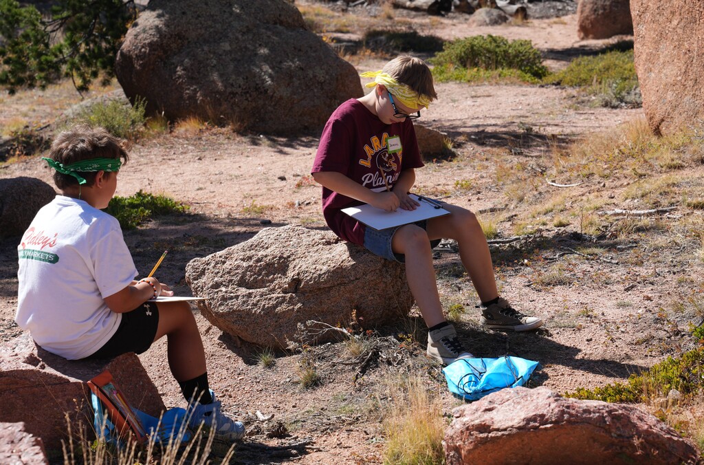 Two boys sitting on rocks drawing on clipboards