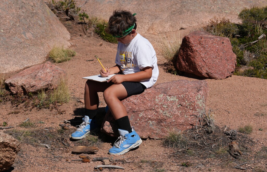 Boy in white shirt and bandana writing on clipboard while sitting on a rock