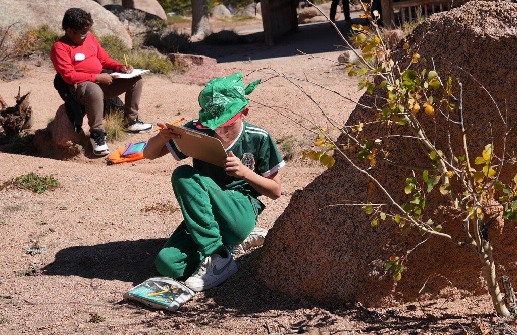Boy in a green shirt kneeling by a rock drawing on a clipboard