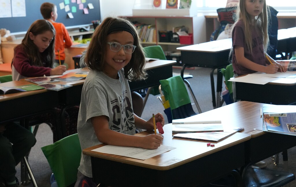 student in blue shirt standing by a desk smiling at camera