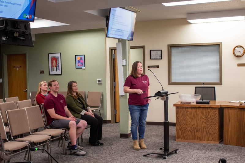 Female with brown hair presenting in front of podium as peers observe