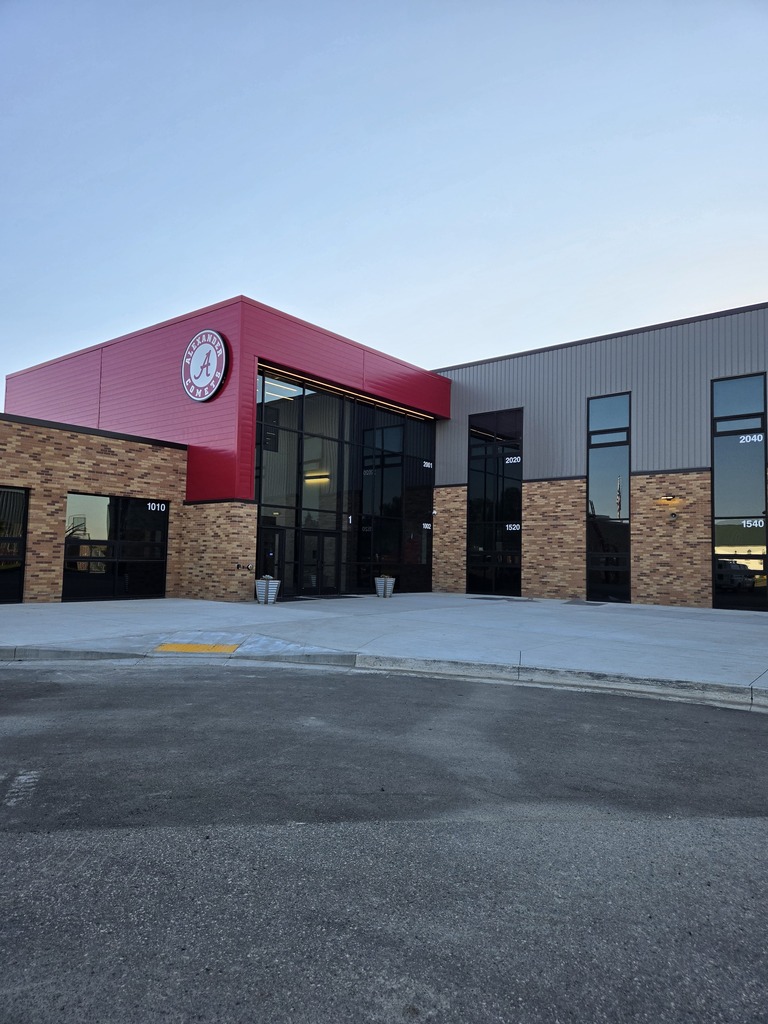 Main entrance of Alexander Public School featuring a red and gray exterior, large glass entryway, brick facade, and the Alexander Comets logo above the entrance.