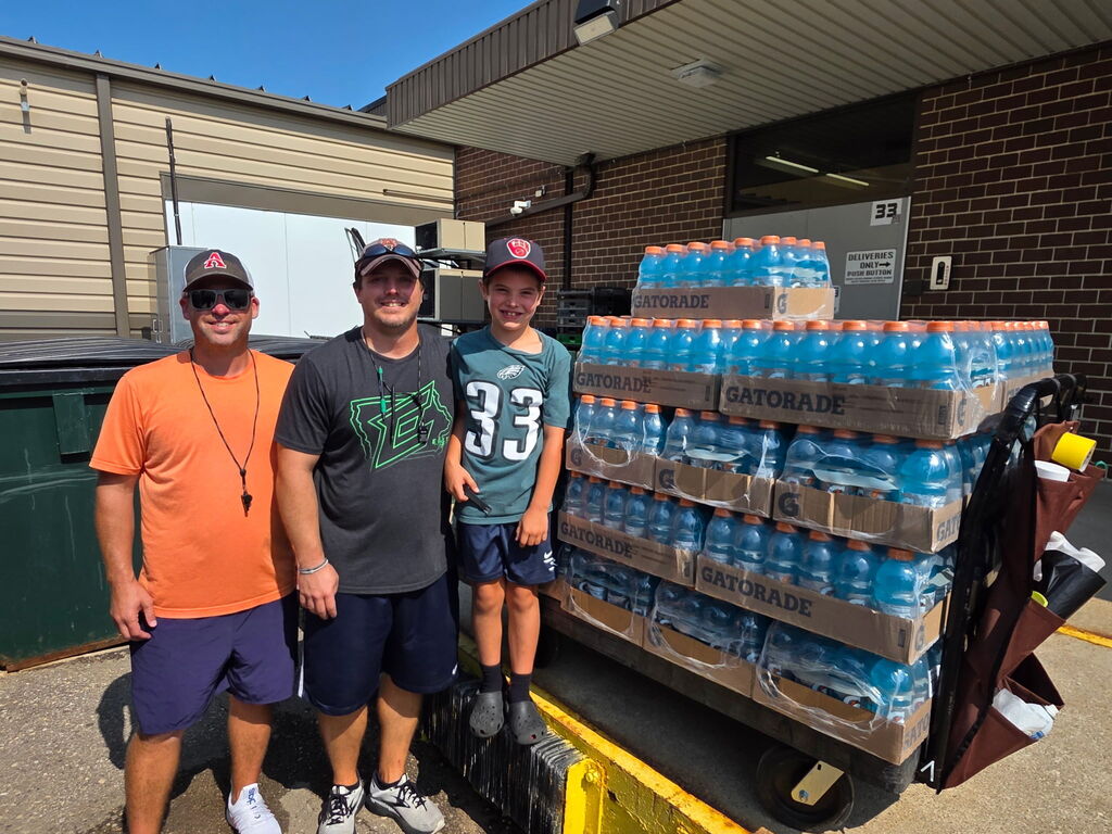 Three individuals pose next to a large cart full of water bottles outdoors in front of a building.