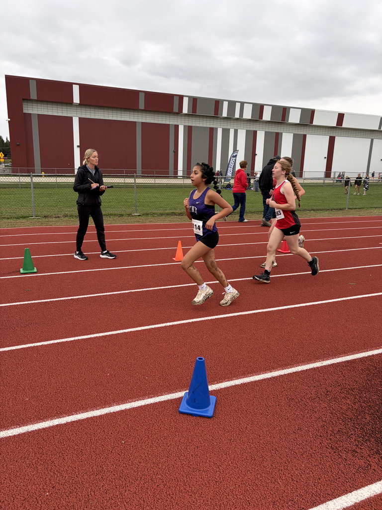 Runners on a track with red lanes, one leading, an official on the sideline, and a large building in the background.