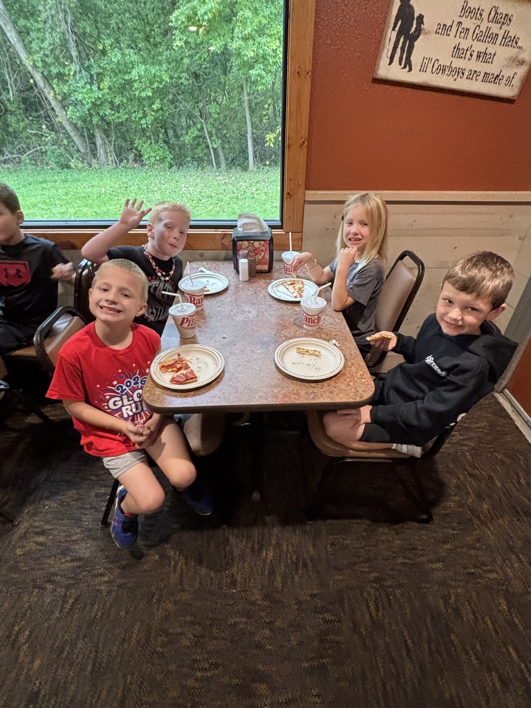 Six children seated around a table. One child has a piece of pizza on his plate. The table has a sign on the wall behind it.
