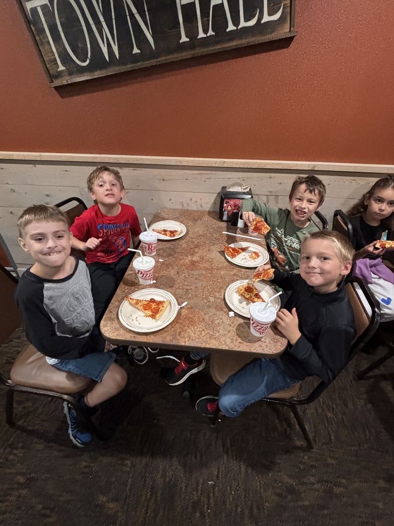 Six children seated around a table with plates of pizza and cups, a wooden sign reading "Town Hall" above.