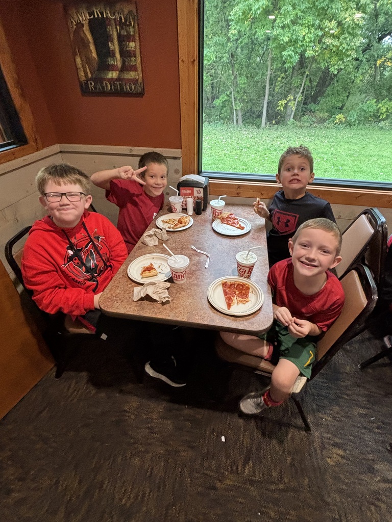 Four children seated at a restaurant table, each with a slice of pizza, smiling. A window with trees outside in the background.
