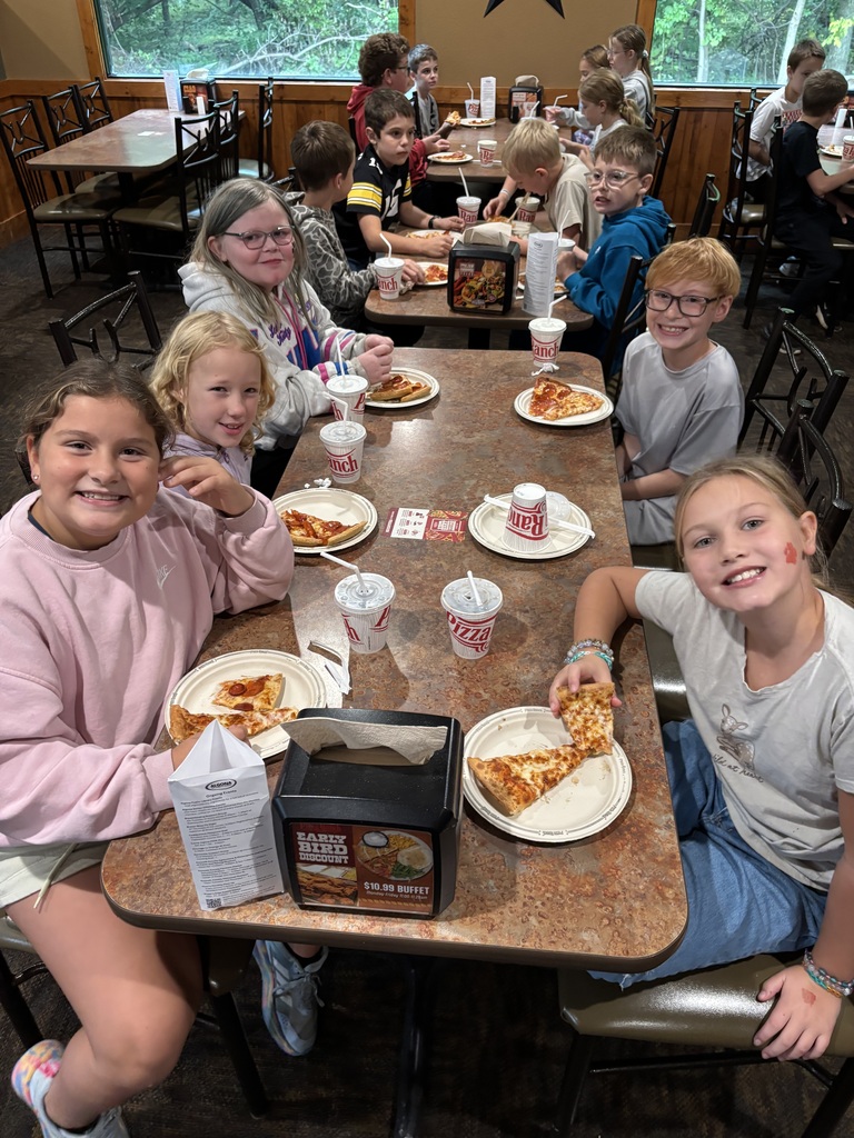 A group of kids sit at a table with pizza slices, cups, and napkins, inside a dining area.