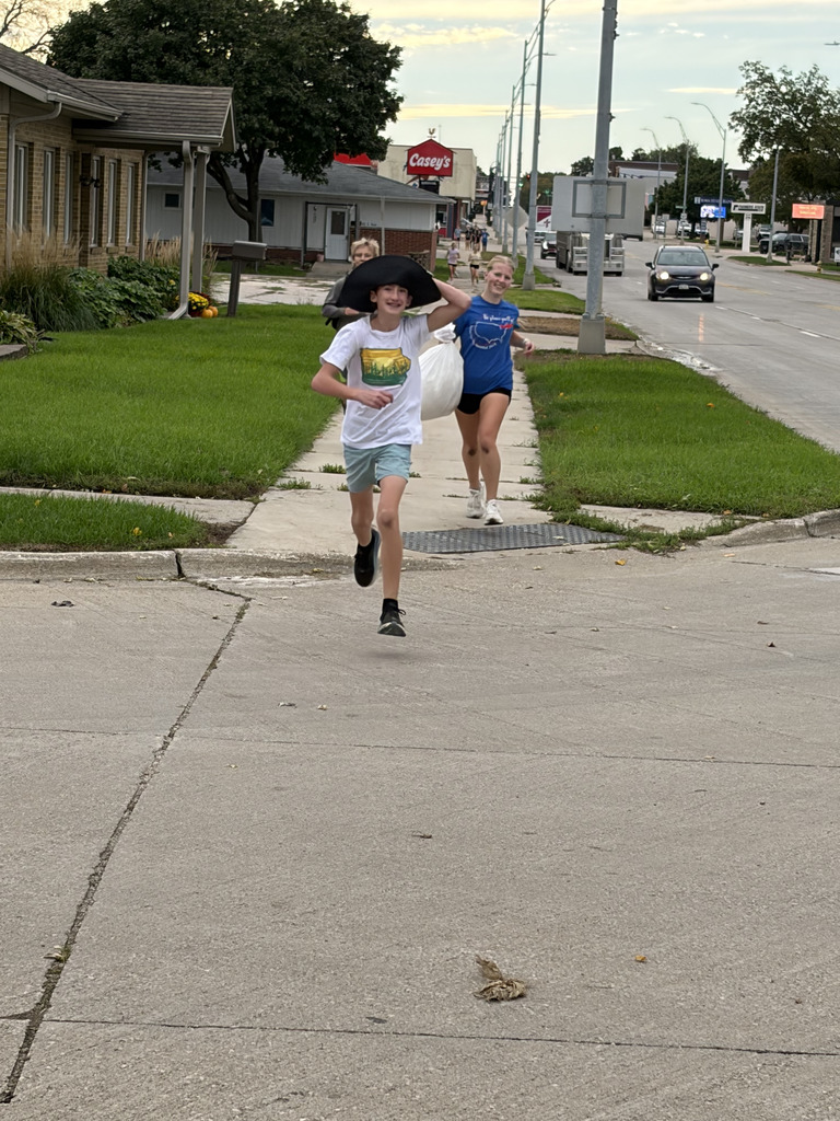 Two children run on a sidewalk, one carrying a hat and the other a bag. The road is visible with a car passing by.
