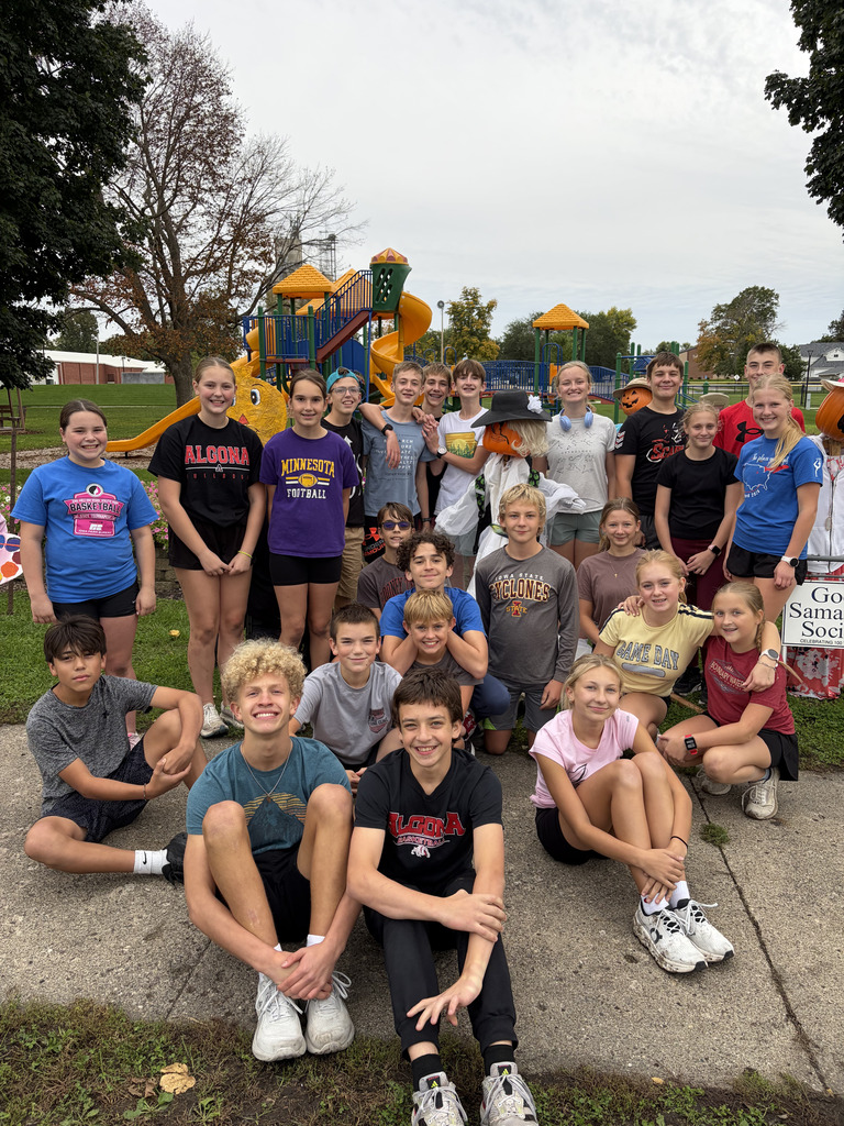 Group of children and adults sitting and standing in front of a playground with a sign.