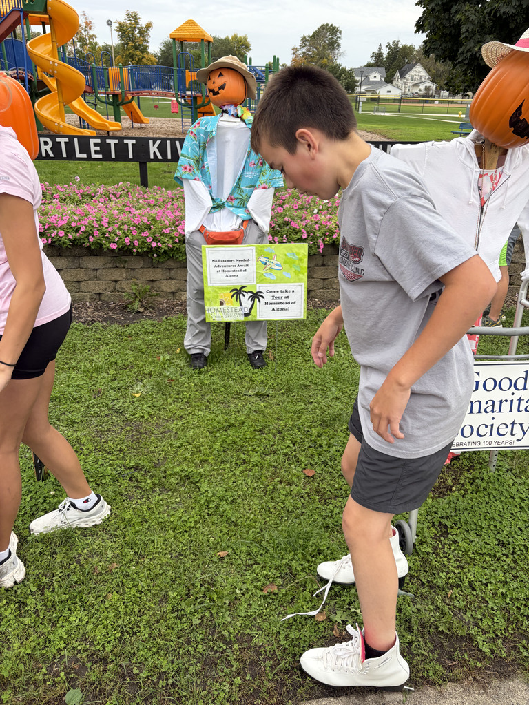 A boy with a pumpkin hat stands on a grassy field. Behind him, another person with a hat is by a sign.