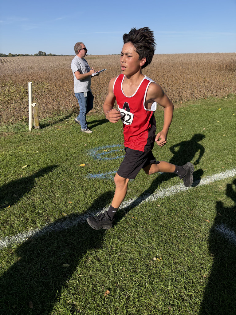 A young boy in a red and black jersey with number 22 sprints along a grassy track. A man stands behind him, observing.